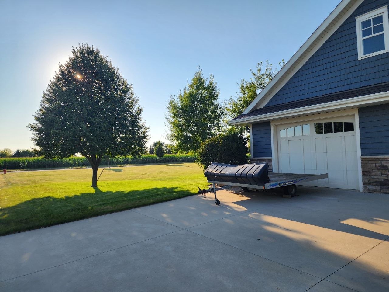 A mowed green lawn beside a blue two-story home, with a large maple tree and a cornfield behind it.