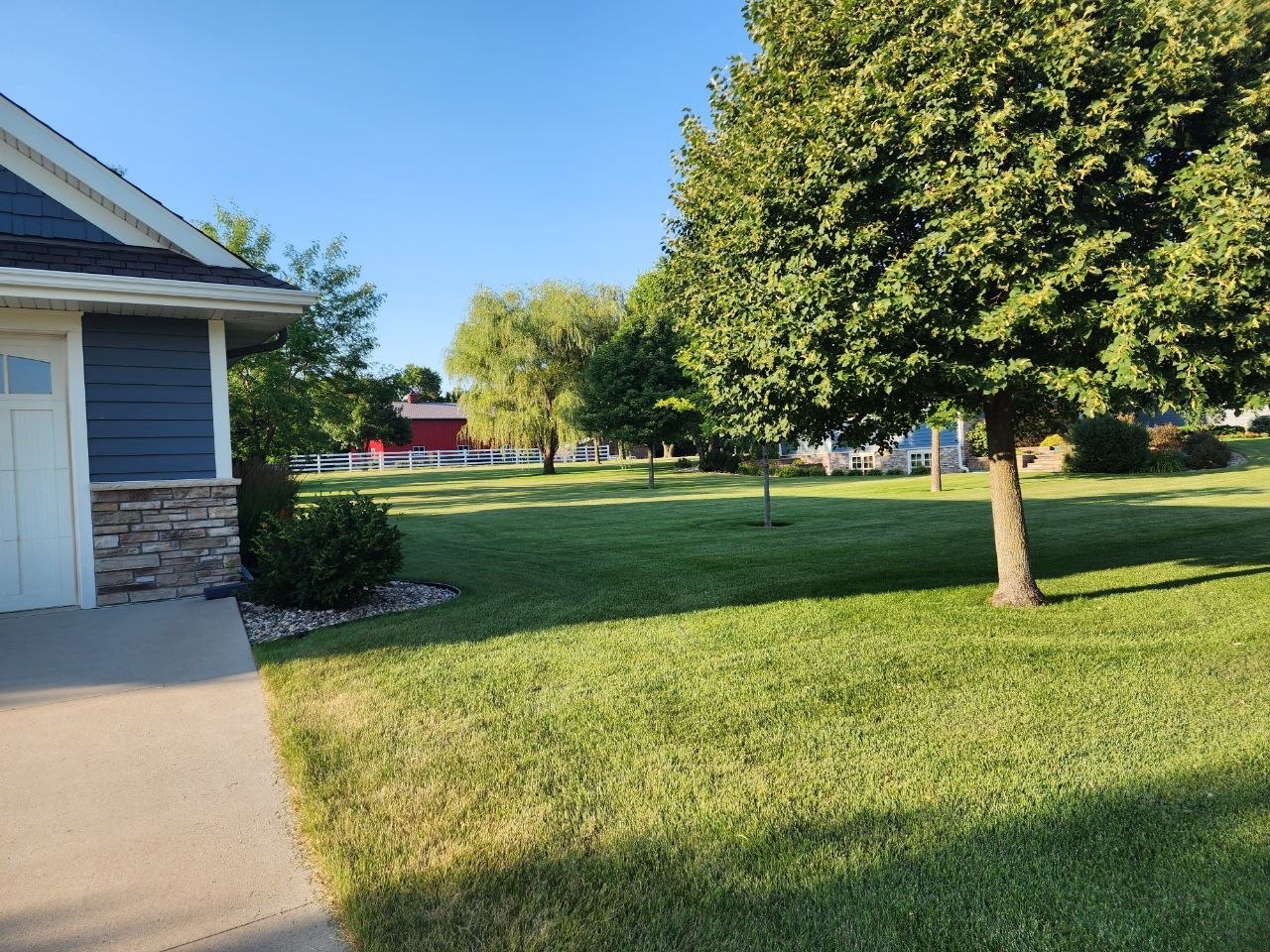 A wide, freshly mowed green lawn with mature shade trees beside a home, a white rail fence and a red barn in the distance.
